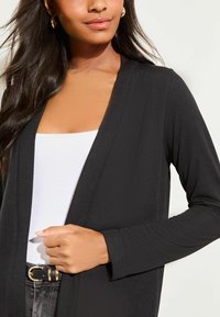 Fekete, hosszú ujjú kardigán rugalmas anyagból, nyitott elülső résszel, fehér trikó fölött viselve. Sima textúrával és finom fényességgel rendelkezik.