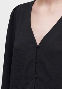 Blouse noire texturée à manches longues avec trois boutons recouverts de tissu et un décolleté en V sur un teint clair.