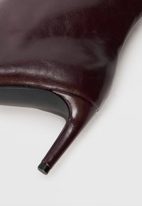 Gros plan sur une bottine en cuir bordeaux foncé à talon aiguille fin sur un fond blanc.