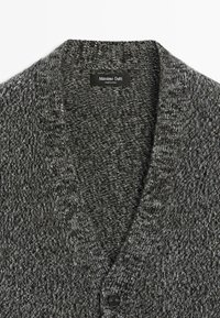 Grå stickad kofta med V-ringning, strukturerat tyg och en svart knapp framtill. Etikett synlig på insidan av kragen.