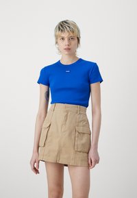 T-shirt blu a coste con colletto rotondo e piccolo logo; abbinato a una gonna color cammello con tasche realizzata in tessuto leggero.