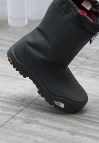 Bota aislante negra con material texturizado, diseño alto y suela de goma con un patrón de relieve. El logo es visible en el costado.