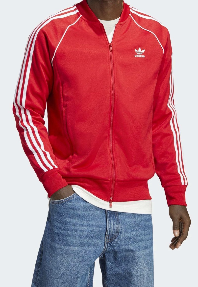 Červená sportovní bunda z hladké látky s předním zipem, bílými třemi proužky na rukávech a malým logem na hrudi.