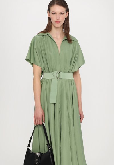 Vestido verde com colarinho, de mangas curtas e cinto ajustado na cintura. O tecido é liso e fluido, combinado com uma bolsa preta.