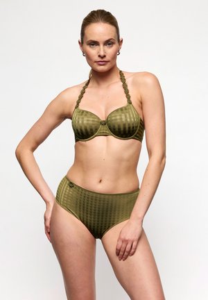 Mujer de piel clara y cabello recogido lleva un sujetador verde a cuadros y ropa interior de talle alto, posando con una mano en la cadera sobre un fondo blanco.