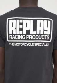 Μαύρο μπλουζάκι με μεγάλα λευκά γράμματα: "REPLAY RACING PRODUCTS" και "Ο ΕΙΔΙΚΟΣ ΣΤΙΣ ΜΟΤΟΣΙΚΛΕΤΕΣ" σε έντονη, σύγχρονη γραμματοσειρά.
