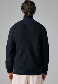 Marineblauwe gebreide trui met hoge kraag, geribbelde structuur en lange mouwen. Voorzien van een aangesloten tailleband en manchetten voor een snug fit.