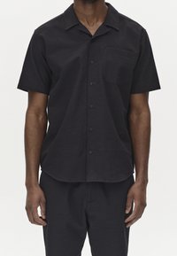 Mężczyzna w czarnej, krótkodziełkowej koszuli zapinanej na guziki z kieszenią na piersi oraz dopasowanych czarnych spodniach na jednolitym tle.