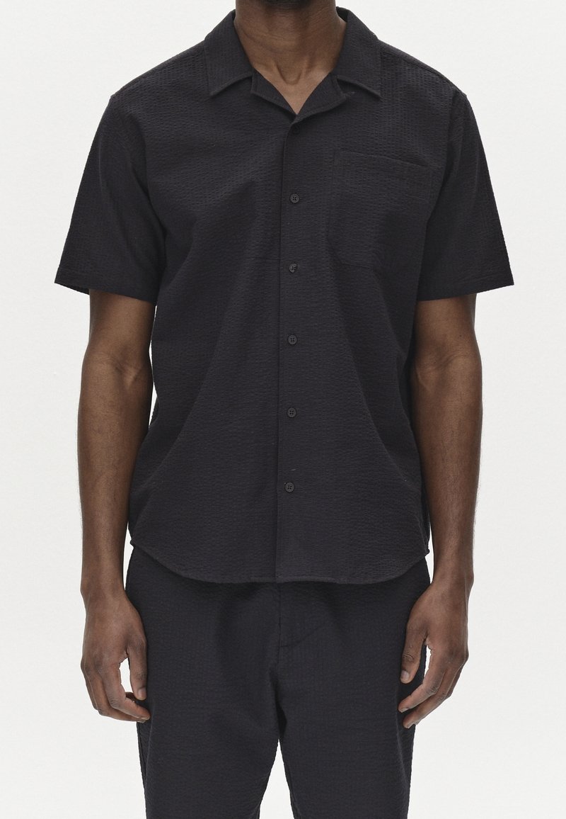 Mężczyzna w czarnej, krótkodziełkowej koszuli zapinanej na guziki z kieszenią na piersi oraz dopasowanych czarnych spodniach na jednolitym tle.