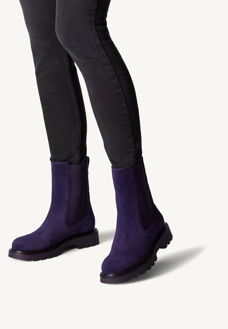 Bottes de cheville en suédine violette avec des panneaux latéraux élastiques et une semelle robuste, associées à un jean skinny noir, mettant en valeur un design épuré.