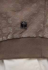 Brązowy sweter z kwiatowym wzorem, o teksturowanej powierzchni, żebrowanym dołem i metalicznym logo w kształcie kwadratu.