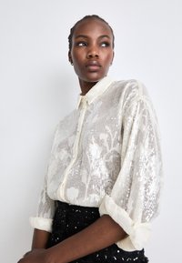 Prozirna, off-white bluza s dugim rukavima, s uzorkom cvjetova od šljokica, rolane rukave i ovratnik. Crna suknja sa šljokicama vidljiva na struku.