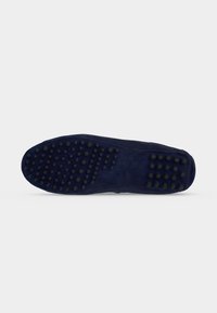Semelle de mocassin en daim bleu foncé avec des crampons ronds noirs en caoutchouc espacés régulièrement au niveau du talon, de la voute plantaire et des orteils sur fond blanc.