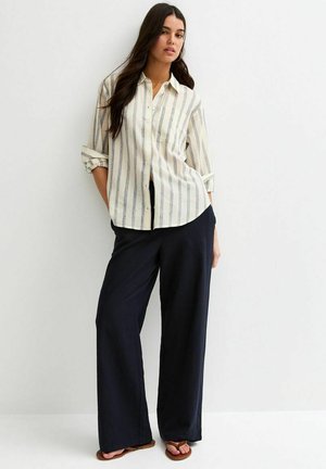 Femme portant une chemise boutonnée à rayures beige et grises avec les manches retroussées, un pantalon noir ample et des sandales marron, debout contre un mur blanc.