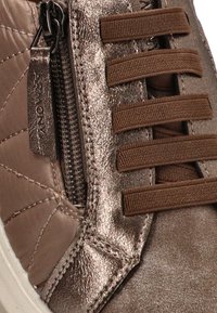 Acercamiento a una zapatilla marrón con panel lateral metálico texturizado, cordones elásticos marrones y cremallera lateral con tirador de marca.