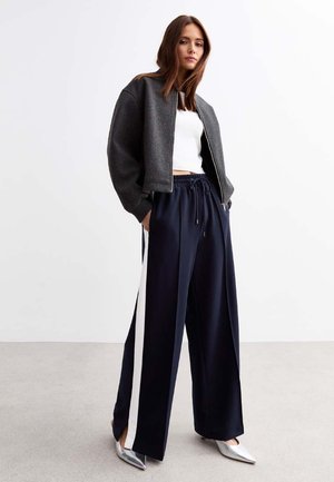 Giacca bomber grigia corta sopra una maglietta bianca, abbinata a pantaloni larghi blu navy con strisce laterali bianche. Scarpe argentate a punta completano l'outfit.