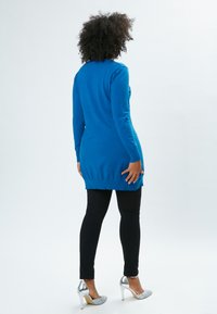 Maglione tunica blu a maniche lunghe con orlo arrotondato, abbinato a leggings neri e scarpe con tacco alto argentate. Texture complessivamente liscia.