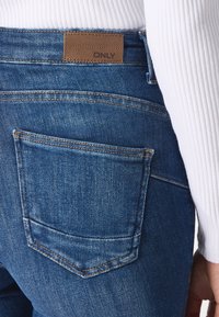 Blå denimjeans med en texturerad brun etikett vid midjan och synlig sömnad. Bakfickan har en böjd design och klassisk skärning.