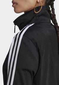 Svart jacka med dragkedja och tre vita ränder längs axlarna, hög krage och ett strukturerat tyg. Metall-dragkedjedetalj synlig.