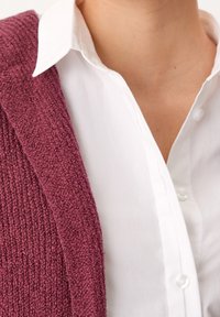 Cardigan bordeaux lavorato a maglia con una finitura testurizzata, abbinato a una camicia bianca impeccabile. Focus sui dettagli del tessuto e sul contrasto di colori.