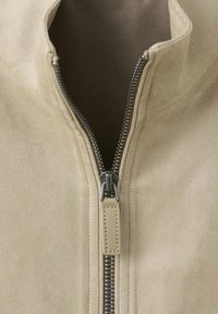 Veste en daim beige avec une fermeture éclair métallique noire partiellement ouverte et un tire-zip en cuir beige assorti.
