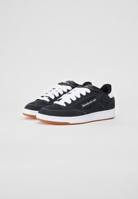 Tênis de camurça pretos com atacadores e costuras brancas; apresenta uma sola de borracha natural, colarinho acolchoado e o logótipo da Reebok na lateral.