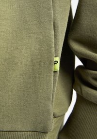 Felpa verde oliva con polsini e orlo a coste, caratterizzata da un piccolo etichetta gialla sulla cucitura, realizzata in morbido tessuto testurizzato.