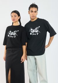 Svarte kortermede t-skjorter har et hvitt grafisk design med Disney-figurer. Den mannlige modellen har på seg lyse jeans, mens den kvinnelige modellen har et svart skjørt.