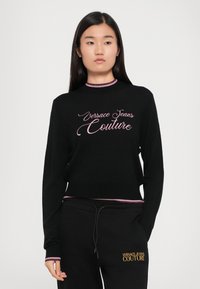 Črn pulover z rožnato vezenim logotipom "Versace Jeans Couture", z rebrastim ovratnikom in manšetami, nosi se z usklajenimi črnimi trening hlačami.