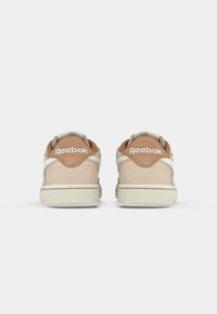 Un paio di sneakers Reebok beige e marroni viste da dietro con suole bianco sporco su uno sfondo bianco uniforme.