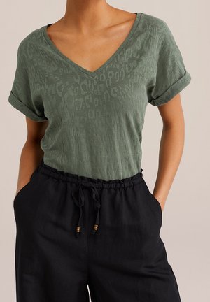 Woman wearing a green short-sleeve V-neck top with rolled cuffs tucked into high-waisted black drawstring pants with hands in pockets.
