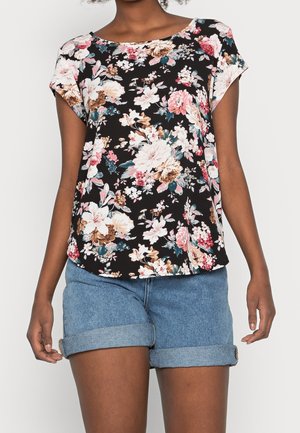 Woman wearing a black floral short-sleeve blouse paired with blue denim rolled-up shorts, standing against a plain light background.