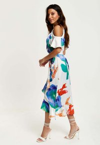 Floral dress in white with vibrant blue, green, and orange patterns. Features ruffled sleeves and a knee-length, flowy skirt. Strappy white heels.