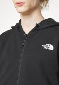 Sweat à capuche zippé noir avec cordons. Présente un logo blanc sur la poitrine gauche et un tissu doux et texturé. Vue de près.