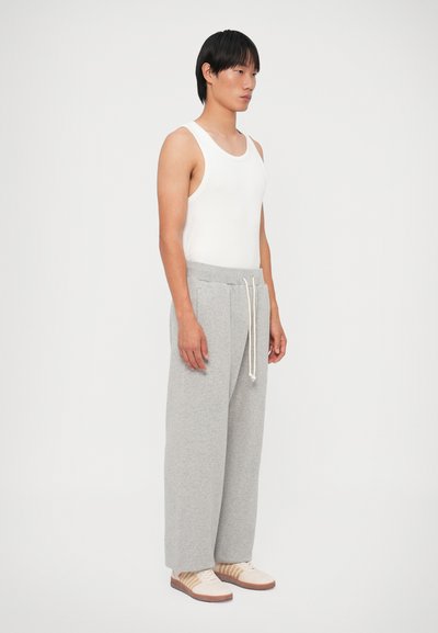 Grijze katoenen sweatpants met een trekkoord in de taille, gecombineerd met een witte tanktop en beige sneakers. Model staat in profiel tegen een witte achtergrond.