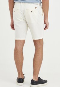 Hvite bomullsshortser med to baklommer og brune knappedetaljer. Brukt med svarte sneakers med hvite såler.