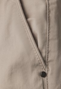 Tissu beige avec une texture lisse et des coutures visibles. Présente un accent en bouton noir sur un design de poche sur mesure.