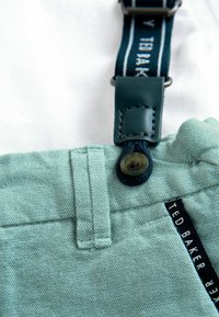 Light teal fabric with a textured finish, featuring a button closure, adjustable navy suspenders with branding, and black label detailing.