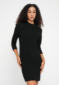 Donna dai capelli ricci che indossa un vestito knit nero aderente a maniche lunghe, in piedi contro uno sfondo bianco.
