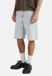 Pantalones cortos de denim azul claro con un ajuste holgado, que presentan piernas rectas, costuras visibles y bolsillos laterales. Llevados con zapatos negros y calcetines blancos.
