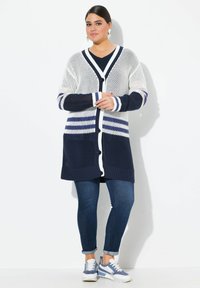 Strikket cardigan i marineblå og hvid, med V-hals, knaplukning og stribet mønster på ærmerne og kanten. Båret over en mørk skjorte.