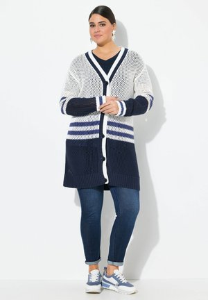 Strickjacke in Marineblau und Weiß, mit V-Ausschnitt, Knopfverschluss und gestreiftem Muster an Ärmeln und Saum. Getragen über einem dunklen Hemd.