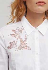 Chemise blanche boutonnée avec broderie en dentelle florale sur le côté gauche, texture de tissu lisse et col classique.