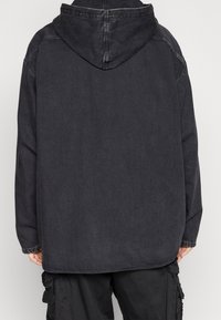 Svart huvad jacka med långa ärmar bärs över svarta cargobyxor, visad bakifrån på en person som står mot en enhetlig bakgrund.