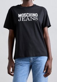 Osoba v černém tričku s nápisem "MO5CH1NO JEANS" vytištěným bílou barvou, v kombinaci se světlými džíny. Pozadí je jednoduché a neutrální.