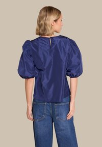 Kvinne med kort blondt hår iført en marineblå bluse med puff-ermer og blå jeans, stående med ryggen til på en beige bakgrunn.