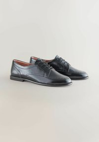 Musta nahka brogue-mekkojalkineet, joissa on nauhalukitus, yksityiskohtainen ompelu ja matalat korot neutraalilla taustalla.