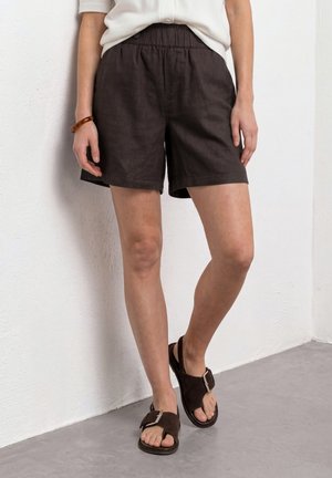 Persoon die zwarte shorts met elastische taille draagt, wit topje, bruine sandalen met gespen, staande tegen een witte gestructureerde muur en grijze vloer.