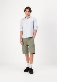 Camisa a rayas azul claro, con mangas largas arremangadas, combinada con pantalones cortos verde olivo. Zapatos negros y calcetines blancos completan el conjunto.
