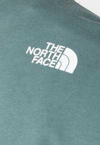 Vitt "The North Face"-logotyp tryckt på texturerad grön tyg, troligen en del av ett plagg som en jacka eller sweatshirt.